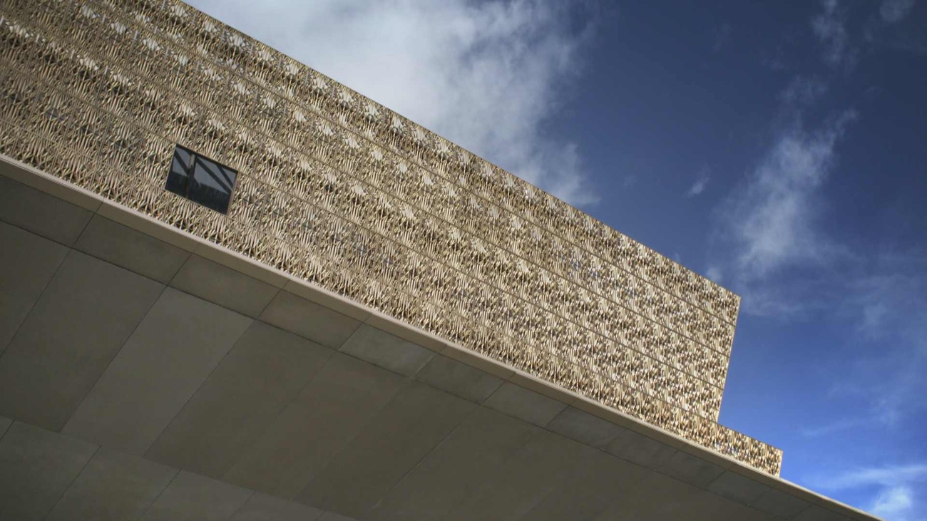 Film Still by Oliver Hardt depicts the structured facade of the National Museum of African American History and Culture. Behind the building is a blue sky.